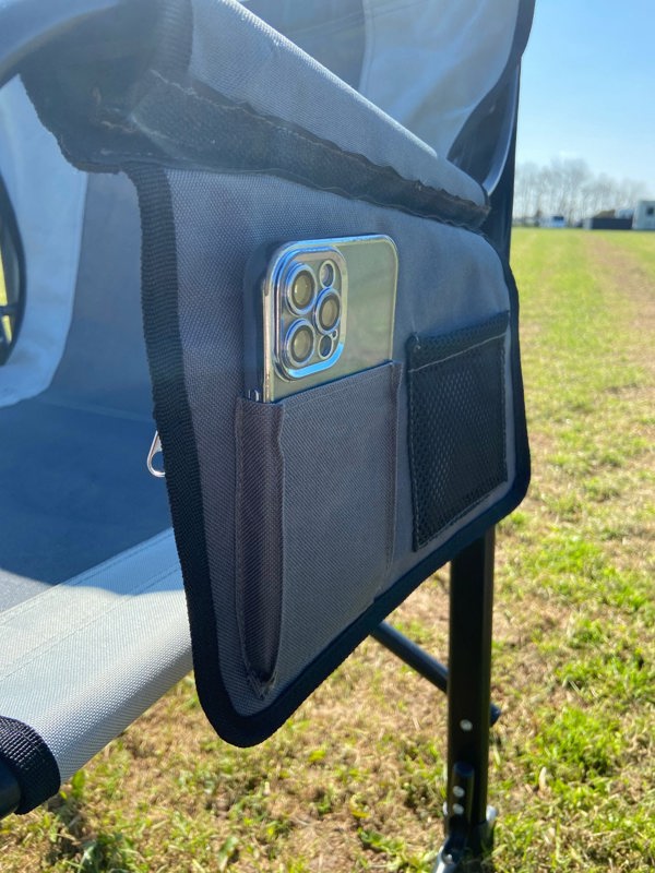 The side storage pocket that contains a mobile phone of the OLPRO Directors Camping Chair in grey being used in a field.