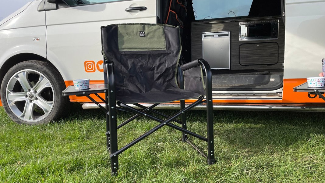 The OLPRO Directors Camping Chair in green and black with the side table up and a melamine mug in the cup holder pictured in front of a VW campervan with its sliding door open.