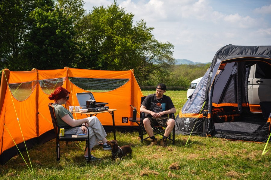 The OLPRO Inflatable Orange Windbreak pitched in a campervan awning set up with an adult female and adult male relaxing on camping chairs.