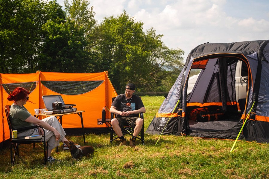 The OLPRO Inflatable Orange Windbreak pitched in a campervan awning set up with an adult female and adult male relaxing on camping chairs.