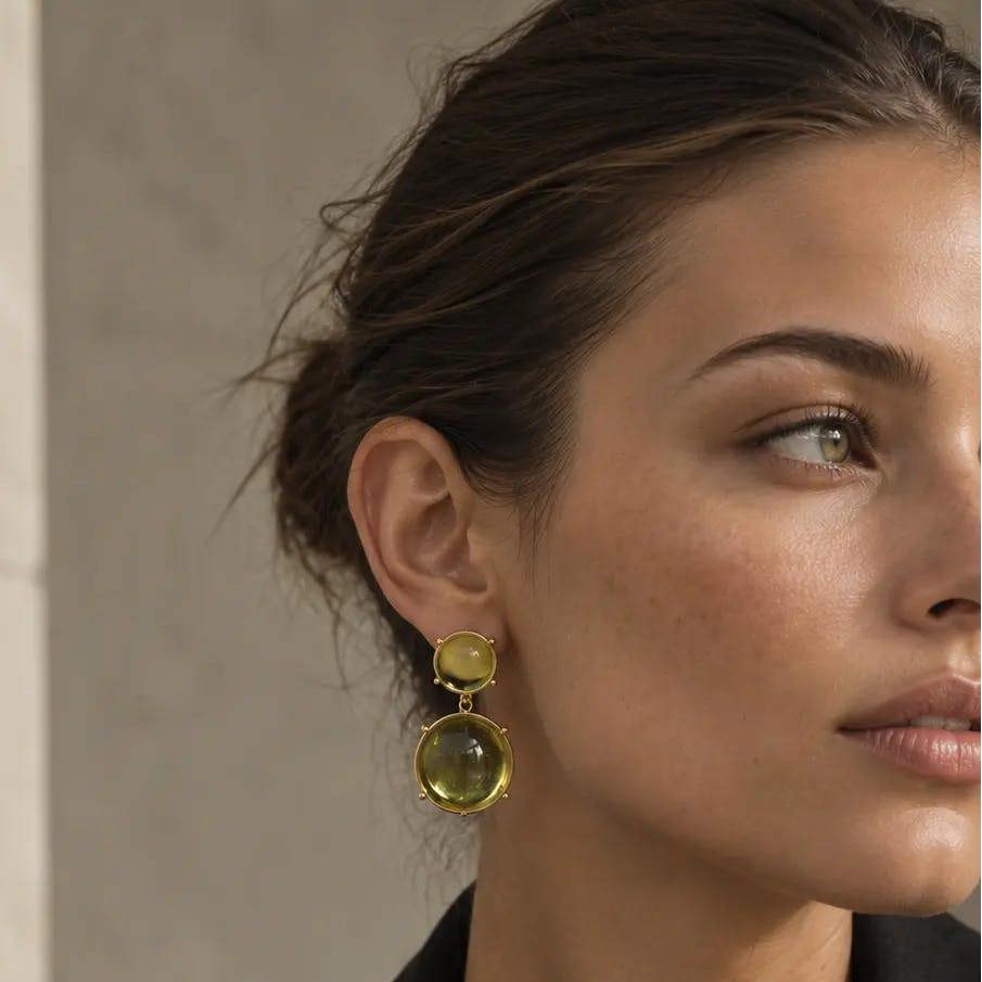 A close-up of a woman wearing a pair of gold drop earrings with round yellow-green gemstones, paired with a black blazer.