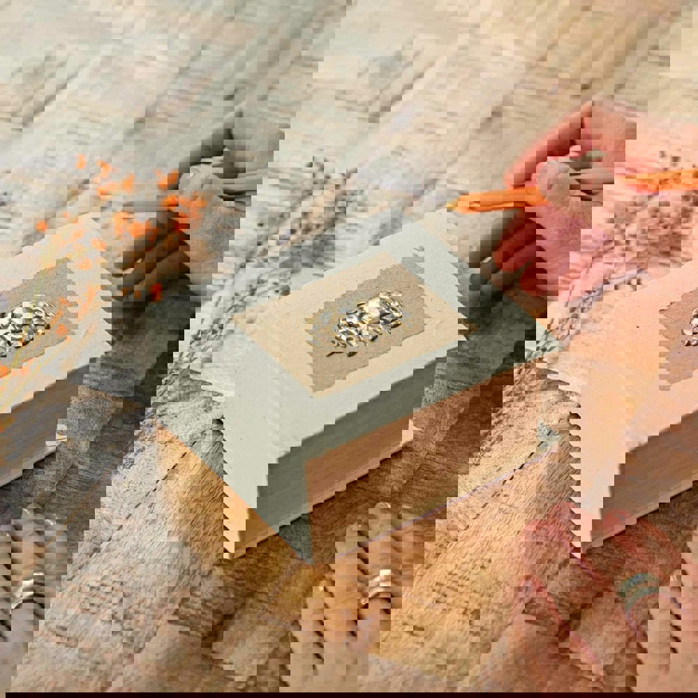 A hand with an orange pen is ready to write in a small, decorative Elephant Dung Memo Block with an embossed elephant on the cover, resting on a wooden table. Nearby are scattered small dried flowers.
