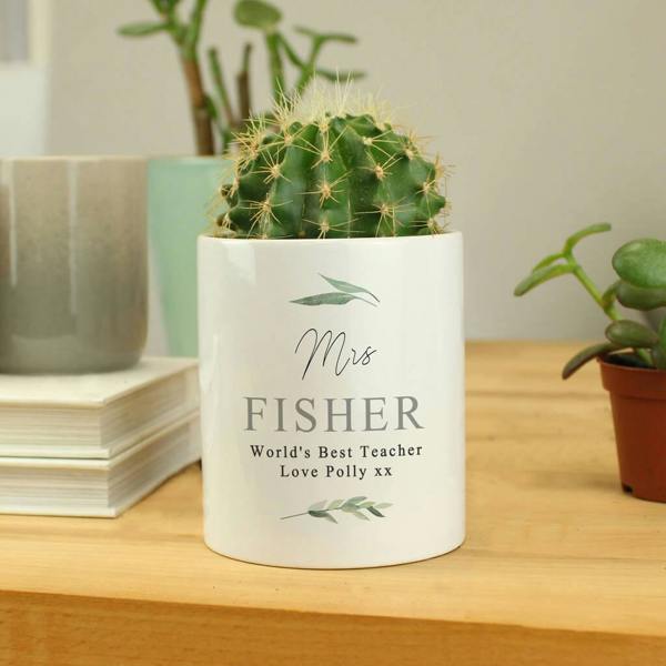 A white ceramic storage pot featuring a botanical leaf design and the text 'Mrs Fisher, World's Best Teacher, Love Polly xx', sitting on a wooden desk with a cactus plant inside.