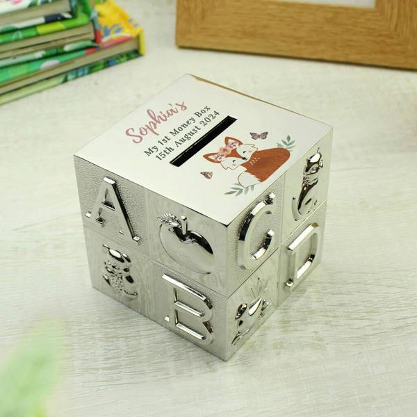 A silver-plated ABC alphabet block money box customized on top with "Sophia's My 1st Money Box 15th August 2024" next to a floral woodland fox illustration, sitting on a light wood desk.