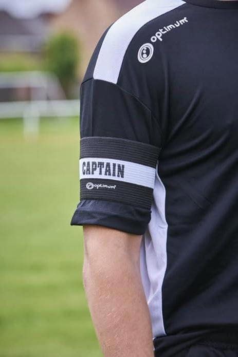 Black Optimum captain's armband worn over the sleeve of a player's black kit on a grass pitch - Optimum