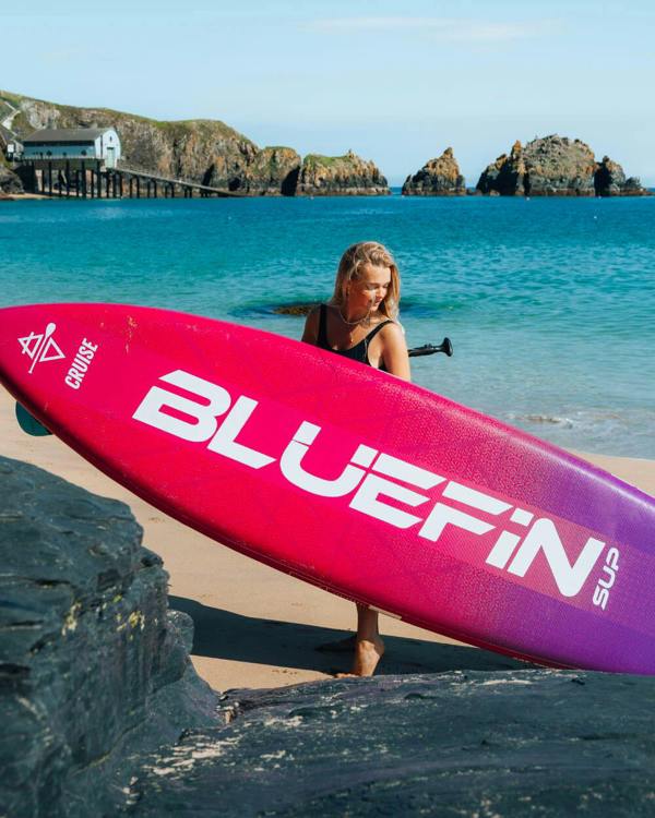 Woman carrying a Bluefin SUP board on a beach with rocks and calm water in the background.