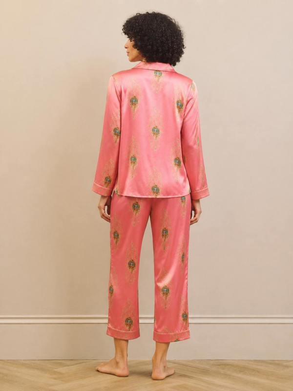 A woman stands barefoot from the back wearing a pink satin pajama set with a small green and gold floral pattern, set against a neutral beige wall and light wood floor.