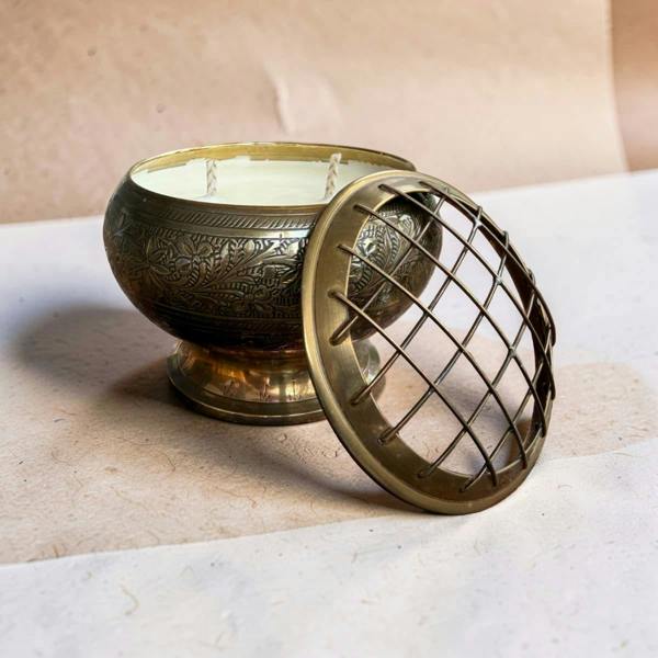 An ornate brass scented candle in a decorative bowl with floral engravings, shown with its matching wire-covered lid leaning against it on a neutral surface.