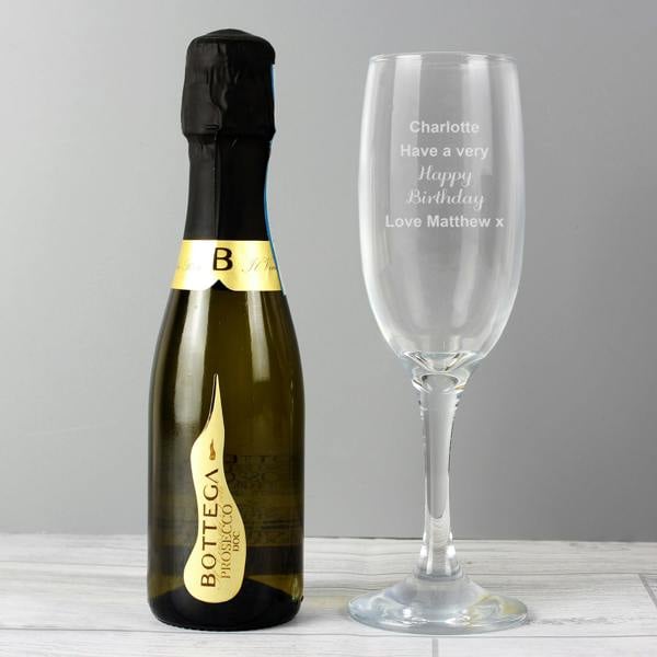 A clear glass champagne flute stands next to a miniature bottle of Bottega Prosecco DOC on a white wooden surface against a grey background. The glass bowl is laser-engraved with the case-sensitive text 'Charlotte, Have a very Happy Birthday, Love Matthew x'.