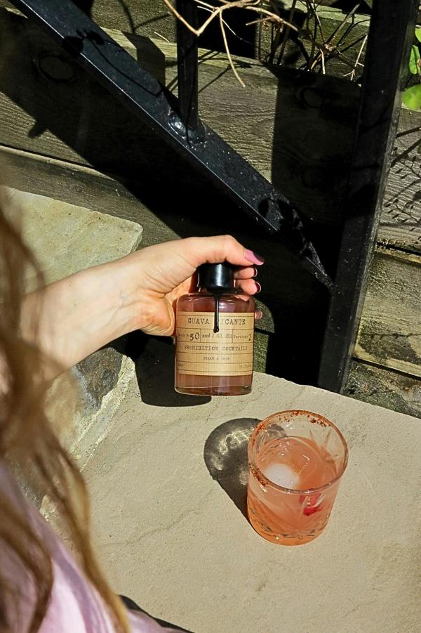 A hand holds a bottle of Guava Picante by Prohibition Cocktails in warm sunlight next to a pink drink on a stone patio.