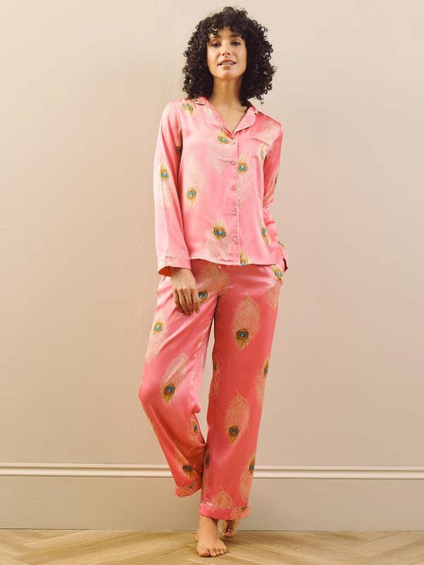 Woman wearing pink pyjama set with peacock feather patterns against a beige wall.