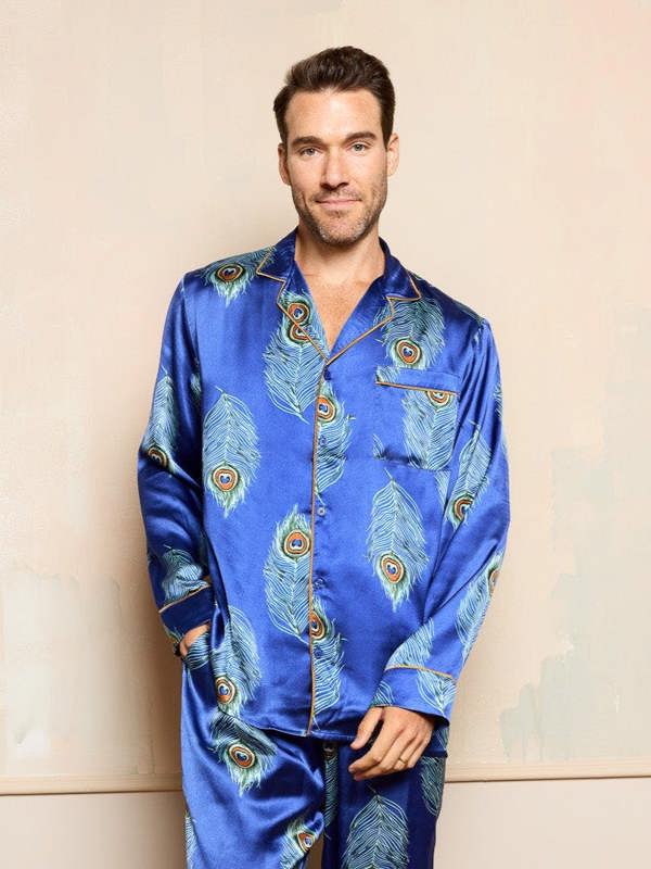 Man wearing a blue satin pyjama set with peacock feather pattern against a beige wall.