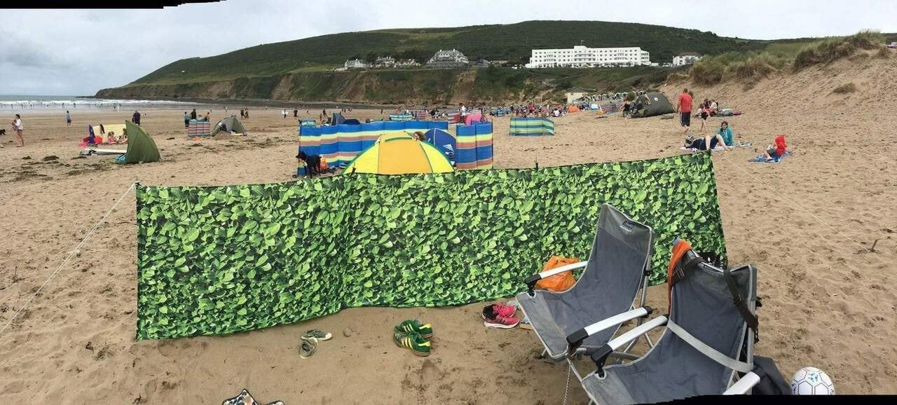 Laurel Hedge 4 Pole Compact Windbreak with Steel Poles by OLPRO pictured on a beach protecting two chairs from the wind.