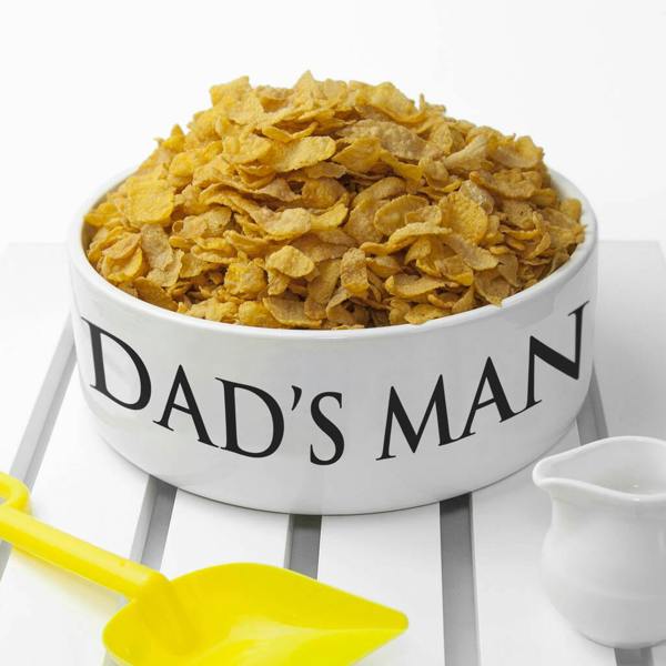 A white, oversized ceramic cereal bowl sitting on a white slatted table, filled to the brim with cornflakes. The words "DAD'S MAN" are printed on the side in large black lettering. A yellow plastic scoop sits on the table next to the bowl.