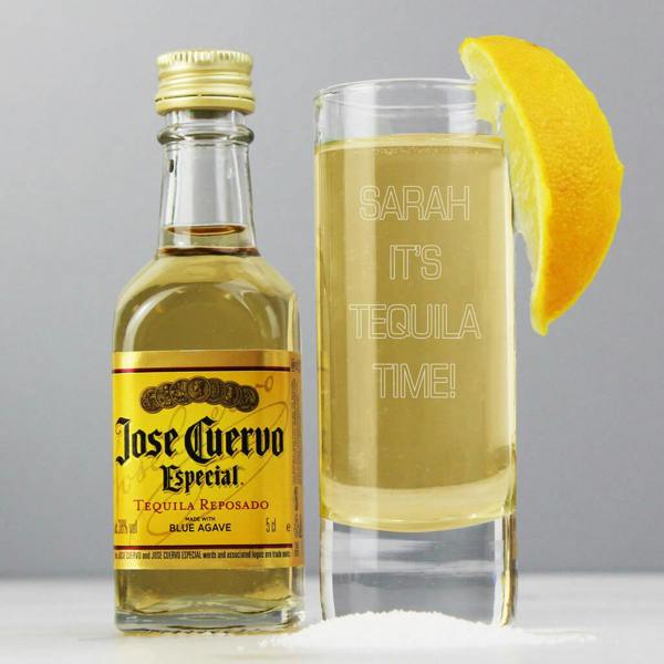 A personalised shot glass engraved with "Sarah It's Tequila Time!" sitting next to a miniature bottle of Jose Cuervo and a lemon wedge.