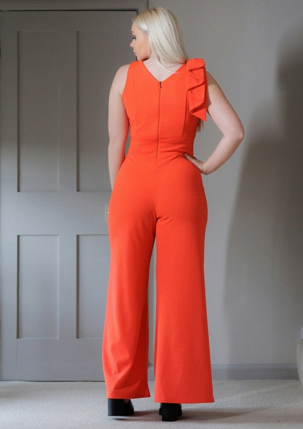 This is the back view of a young lady wearing a Flame red coloured V neck jumpsuit, with wide legs and a bow on one shoulder. She is wearing black open-toed sandals and has her hand on her hip