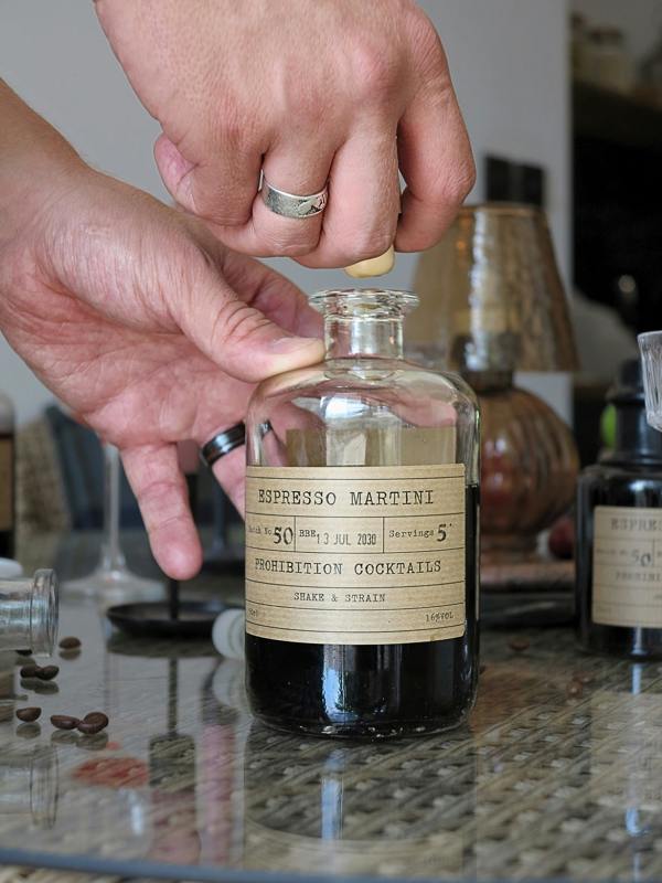 A person's hands unscrew the cap of a vintage-style clear glass bottle labeled 'ESPRESSO MARTINI' and 'PROHIBITION COCKTAILS' on a marble countertop.