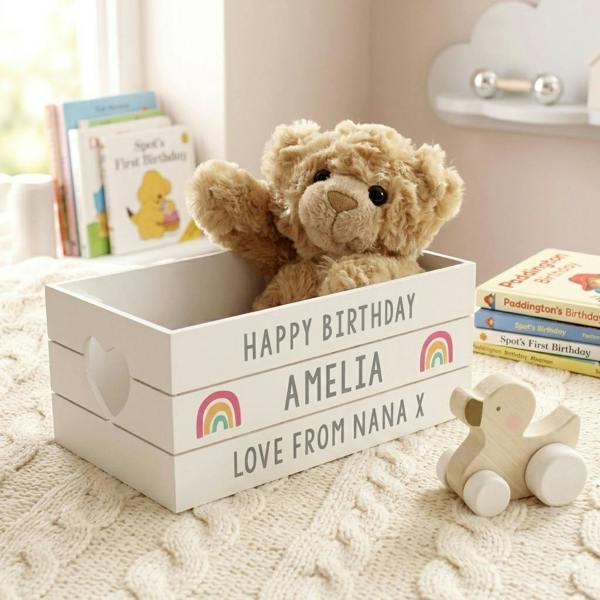 Personalised white wooden birthday keepsake crate with rainbow details, holding a teddy bear on a knitted blanket next to children's books.
