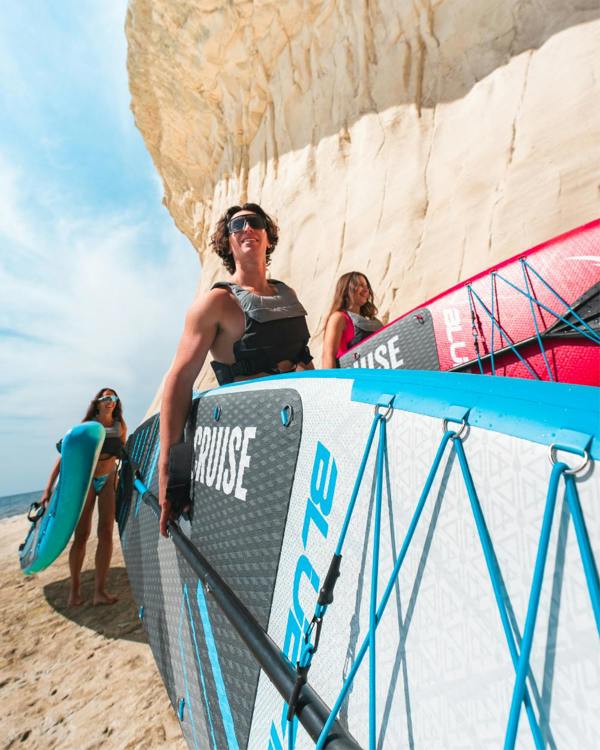 Two people carrying inflatable paddleboards on a sandy beach near a cliff under a blue sky
