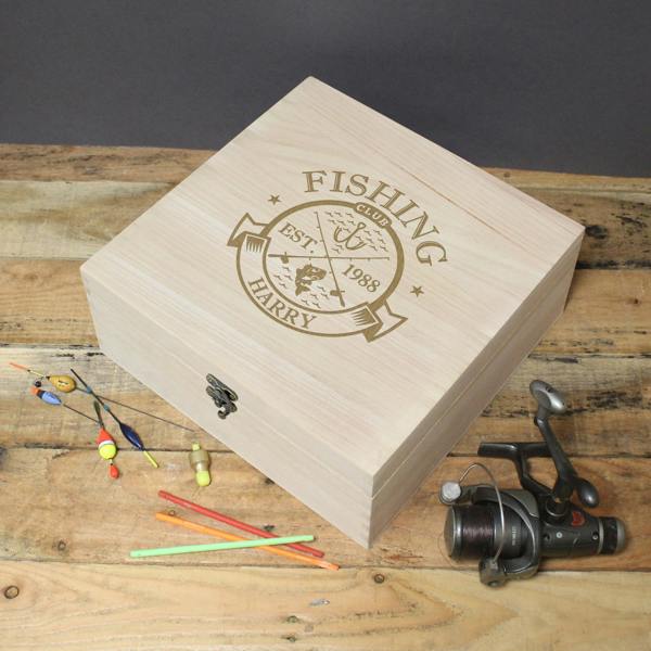 A large, square wooden keepsake box with a metal clasp, featuring an engraved "Fishing Club" logo with a fish and hooks design. The box is personalised with "EST. 1988" and the name "HARRY." It is displayed on a wooden surface next to a fishing reel and several colourful fishing floats.