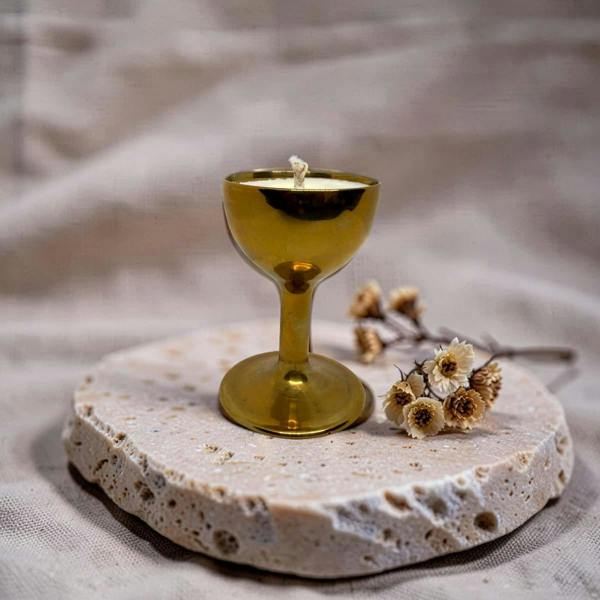 A small gold goblet-shaped candle with a wick sits on a round stone slab, styled with dried flowers against a soft beige fabric backdrop.