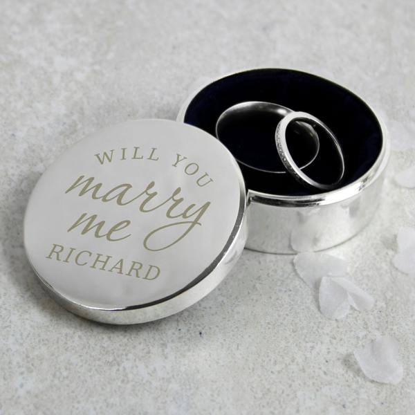 A polished silver round ring box with an open lid engraved with 'WILL YOU marry me RICHARD', styled on a light stone countertop with two silver bands sitting inside the blue velvet interior.