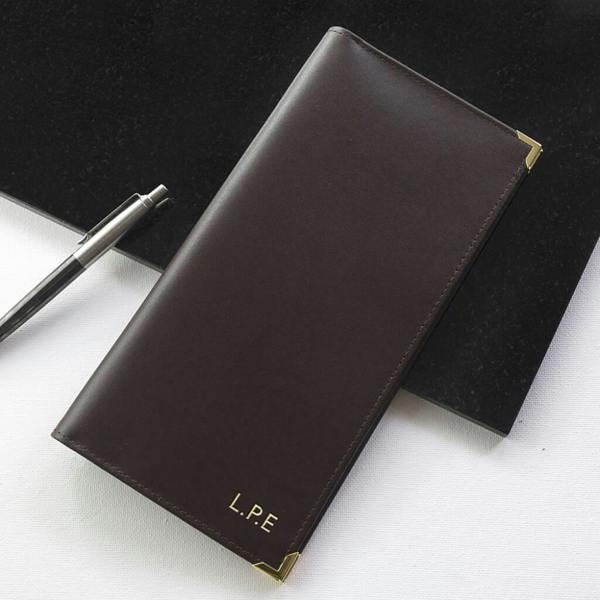 A premium dark brown Italian leather travel wallet with gold corner protectors, embossed with the initials L.P.E in gold lettering, resting on a white surface next to a silver pen.