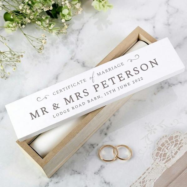 A personalised wooden wedding certificate box with a hand-painted white lid resting on a modern white marble surface alongside two gold wedding rings and gypsophila flowers.
