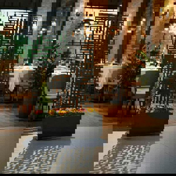 Black metal privacy screen planter with a modern geometric pattern, featuring vibrant flowers blooming inside the base reservoir in a restaurant setting.