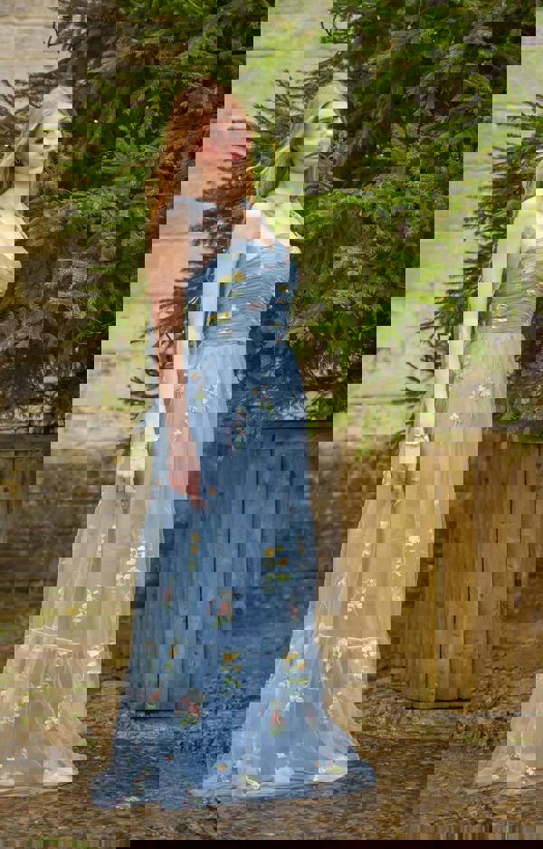 Woman in a blue floral dress standing outdoors with greenery in the background