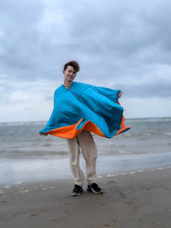 A man along the beach wearing a blue/Orange reversible Blanko as a poncho, with the blue side visible.