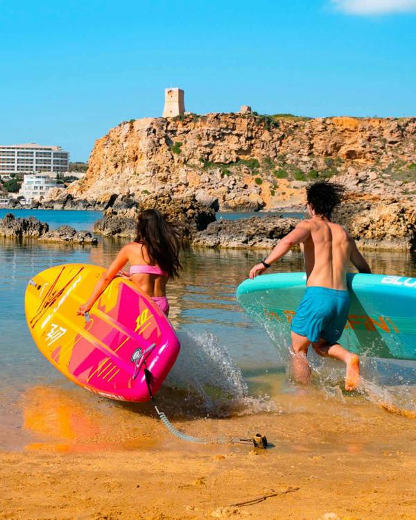 Man and Woman running into the water holding a Blue Lagoon and Pink Coral 10' Lite Bluefin sup paddle board 