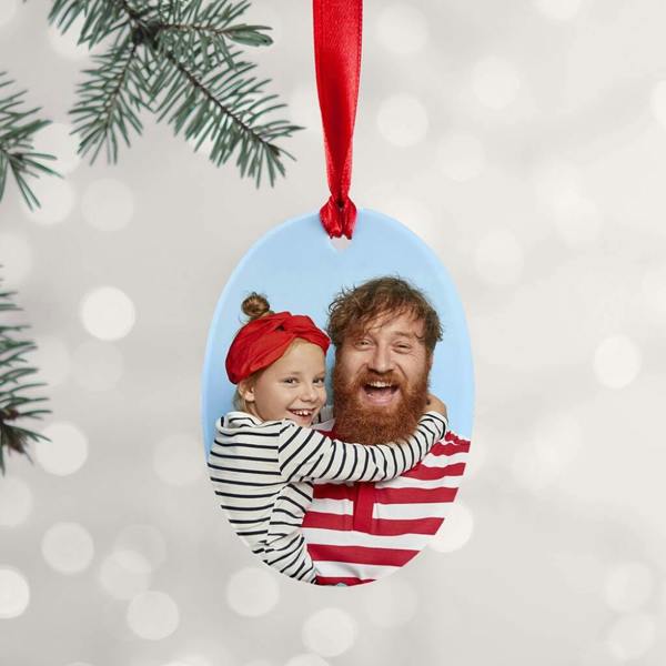 Oval-shaped ceramic personalised Christmas ornament featuring a high-quality photo print of a smiling father and daughter hanging from a tree branch by a red ribbon.