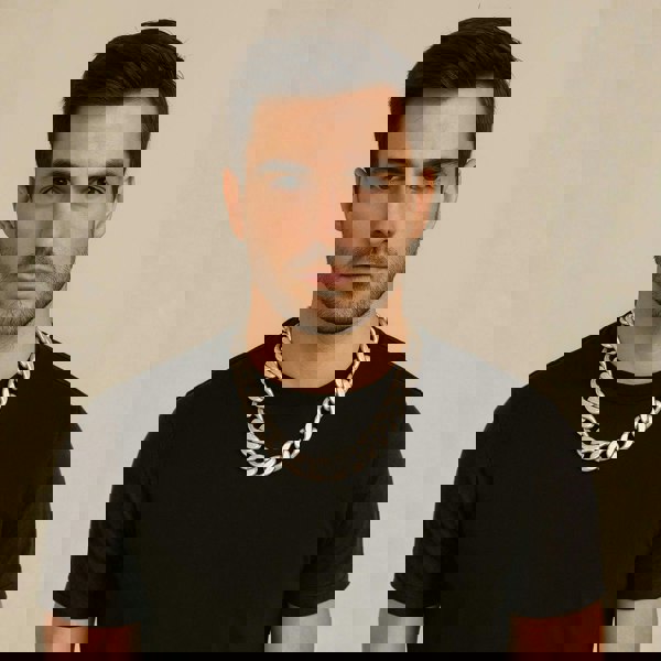 Man wearing a black t-shirt and large silver chain necklace against a beige background