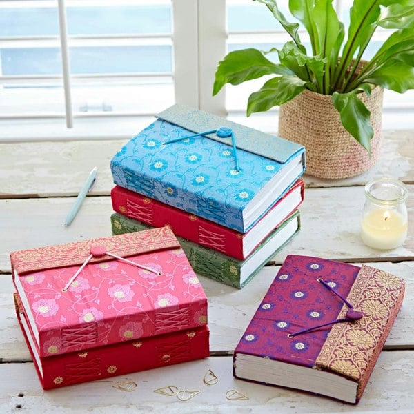 Large Sari Fabric Journals with decorative closures are stacked on a wooden table beside a green plant, candle, and pen.