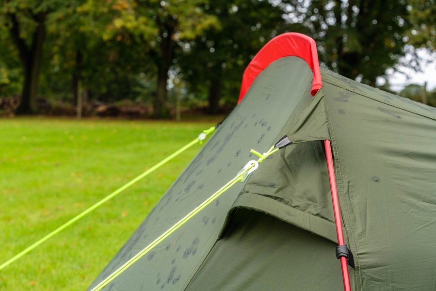 A close up of the pole, pole sleeve, guy ropes and vent of the OLPRO Beckford Lightweight 2 Person Tent (Ripstop) with it being pitched in a field with tress in the background.