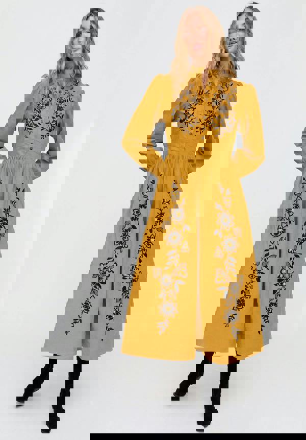 Woman wearing a mustard yellow dress with black floral embroidery on a white background