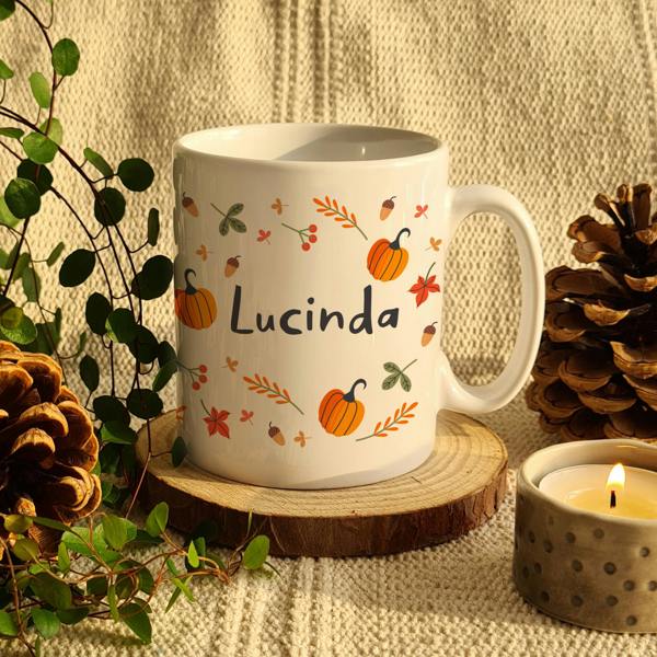 White ceramic mug printed with pumpkins, acorns, and the name Lucinda sitting on a wooden coaster with pinecones and a lit candle