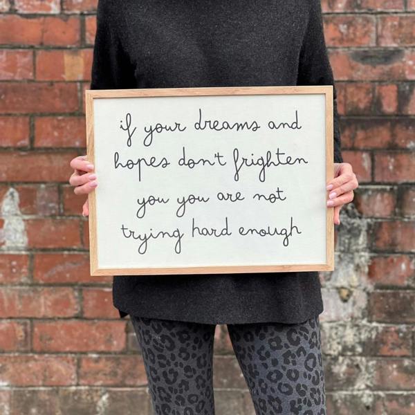 A woman wearing leopard print leggings holding a landscape-oriented solid oak framed white cotton typography print displaying the lowercase quote "if your dreams and hopes don't frighten you you are not trying hard enough" against a brick wall.