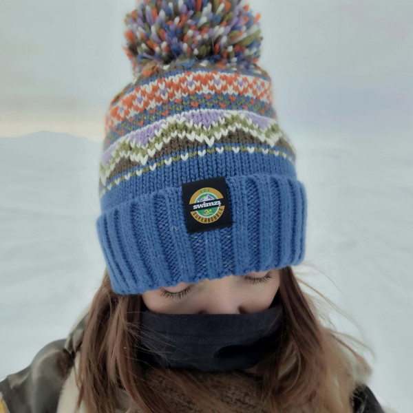 Women wearing a bobble hat with an ocean blue band and farisle knit design in  shades of olive green, lilac, orange and white.  The hat also has a deep ribbed headband and contains reflective yarns and a luxury sherpa fleece lining.