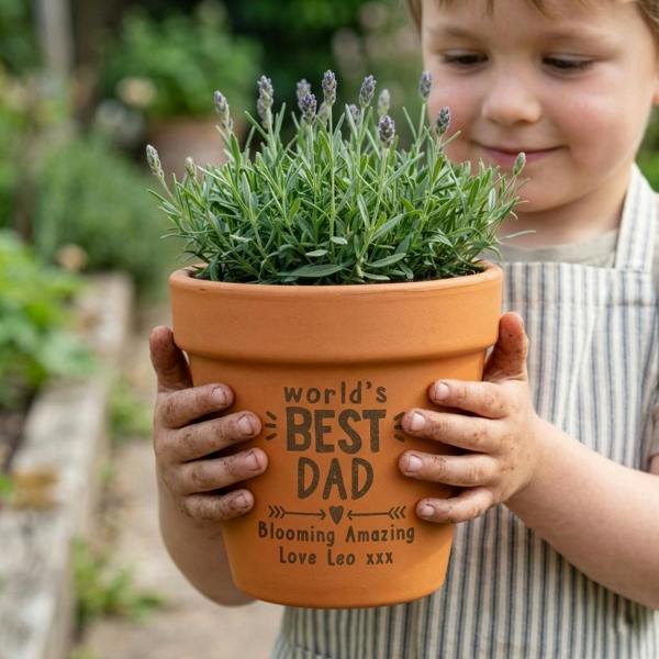 A natural clay terracotta plant pot engraved with "World's Best Dad" holding a fresh green lavender plant on a rustic stone patio, next to small seedlings and gardening gloves.