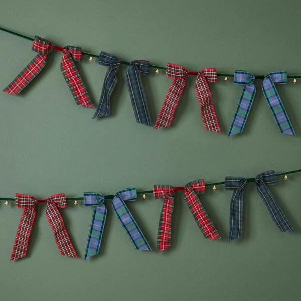 Two parallel rows of a tartan ribbon garland featuring festive plaid bows and tiny gold bells hanging against a matte sage green wall.