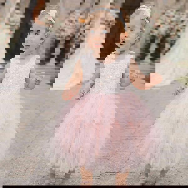 Baby in a pink and white dress standing outdoors with blurred wedding background