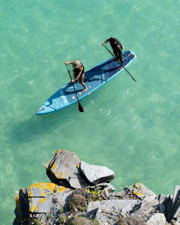 Two paddlers on a blue stand-up paddleboard in clear turquoise water near rocky shore.