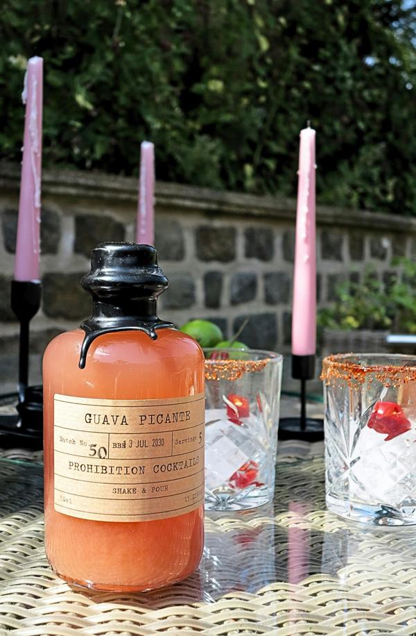 A bottle of Guava Picante by prohibition cocktails with orange liquid, displayed on a woven table alongside two glasses with ice and cherries, set outdoors with pink candles and a stone wall background.