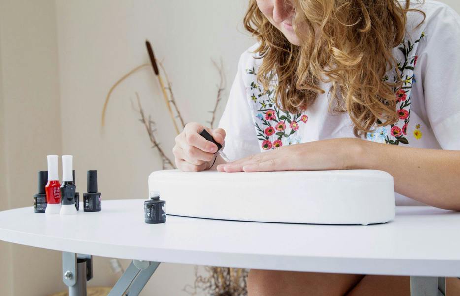 A woman with curly hair paints her nails at a white Rio Professional Manicure & Nail Beauticians Table, surrounded by nail polish bottles. She wears a white top with colorful flower embroidery. The setting is bright and relaxed.