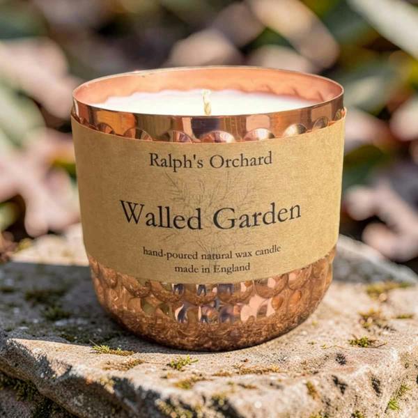 A copper-colored glass candle jar with a hammered finish and a kraft paper label reading 'Ralph's Orchard Walled Garden', photographed outdoors on a stone surface with blurred foliage in the background.