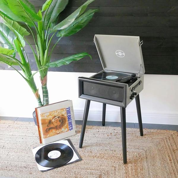 A retro grey vinyl record player on black legs styled in a modern living space next to a large green indoor plant, with a vinyl record on the woven rug below.