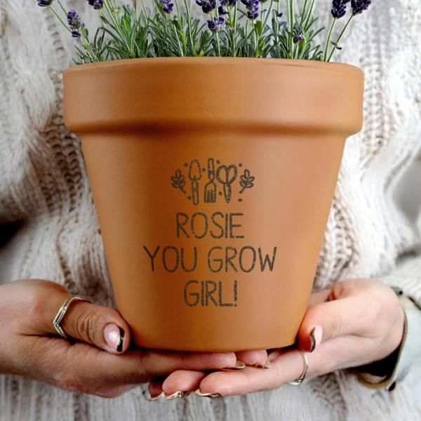 A close-up of a person wearing a white cable knit jumper holding a personalised terracotta plant pot engraved with 'Rosie You Grow Girl' and filled with fresh lavender.