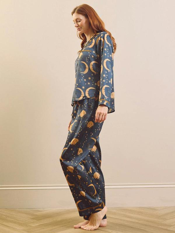 A woman modelling a two-piece pyjama set with a dark blue space-themed print featuring gold crescent moons, stars, and planets, standing against a beige wall and light wood floor.